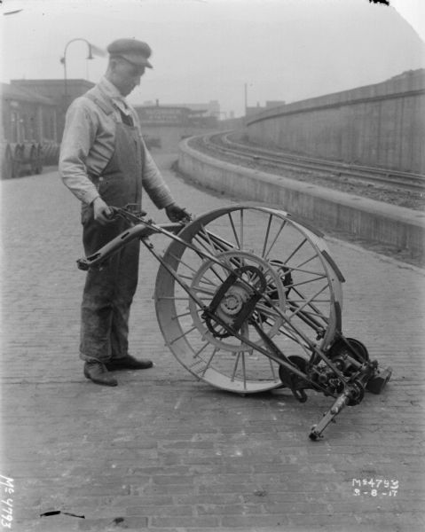 Employee standing and holding an agricultural machinery part, including a large wheel, in factory yard. On the right is an elevated railroad platform, and on the left are factory buildings.