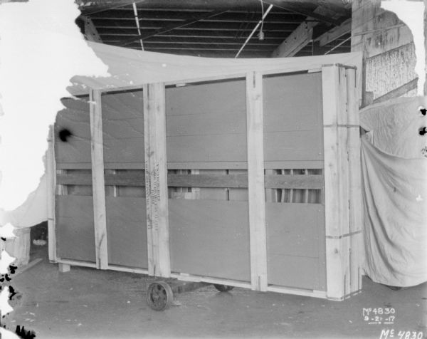 Packing crate with a wagon box inside for machine shipment. White cloth backdrops are set up around the wagon, which is inside a factory.