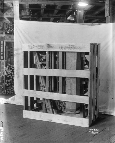 Gun cart crated for shipment on factory floor, labeled "US Machine Gun Cart." There is a backdrop behind the crate, and behind that are materials stored on shelves.