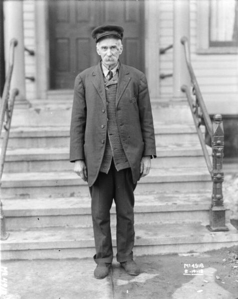 Full-length portrait of a man wearing a coat over a heavy sweater standing outdoors at the bottom of stairs that lead up to a double door.