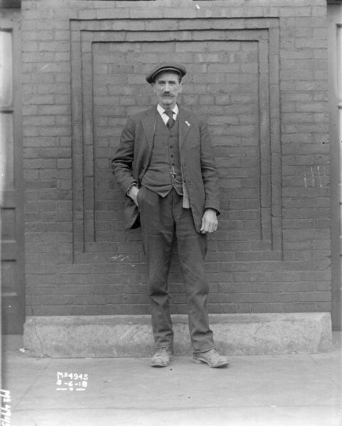 Full-length portrait in front of a brick building of a man in dress clothes, standing with his hand in his pocket.