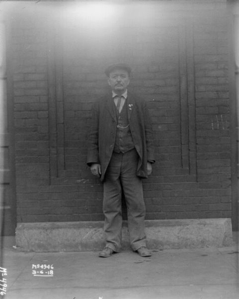 Full-length portrait in front of a brick building of a man in dress clothes.