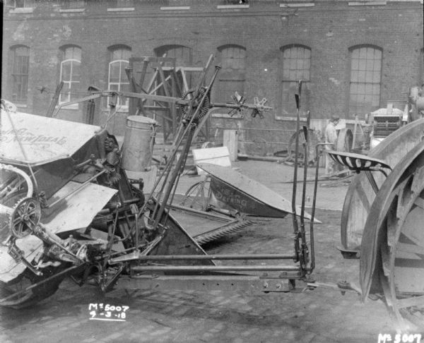 Close-up of a hitch on a tractor-drawn Binder in the factory yard. There is a man in the background near a brick factory building.