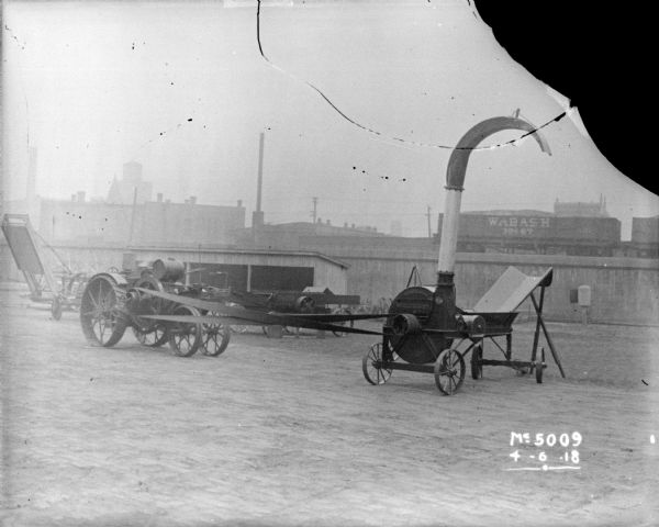 A tractor-powered Ensilage Cutter in the yard at McCormick Works. In the background is a shed, and railroad cars on an elevated railroad platform.