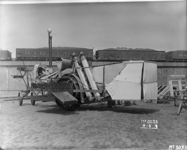 Early model Harvester Thresher in factory yard. In the background are railroad cars on an elevated railroad platform.