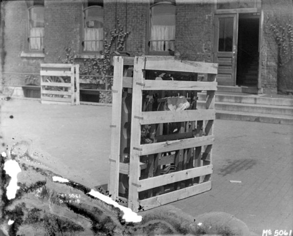 A crate packed with a Gun Cart trench mortar barrel for shipment is outdoors in the factory yard. In the background is a brick factory building.