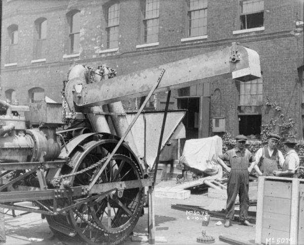 Men are posing with a Harvester Thresher outdoors in factory yard. A brick building is in the background.