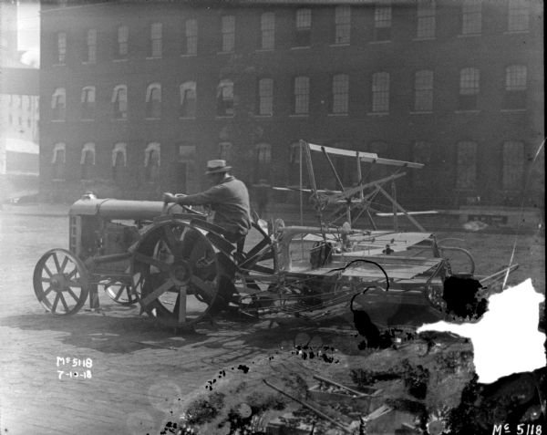 A man is driving a tractor pulling a Binder in factory yard. There is a brick building in the background. 