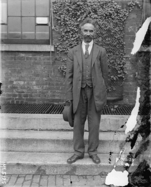 Full-length portrait of a man standing outdoors on steps in front of a brick building. He is wearing a suit and is holding a hat in his right hand.