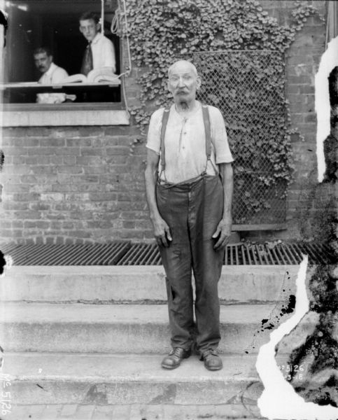 Full-length portrait of a man standing outdoors on steps. He is wearing work clothes. Two men are looking out of an open window from the brick building in the background on the left.