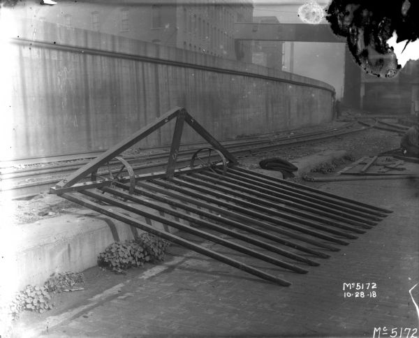 Sweep Rake outdoors in factory yard leaning on concrete near railroad tracks. There is an elevated railroad platform, and factory buildings with a skybridge in the background.