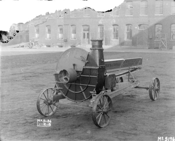 International Ensilage Cutter 'G" in factory yard. An automobile is parked on the left near a brick building is in the background.