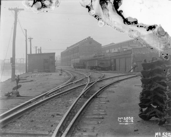 View towards three sets of railroad tracks, each branching off to the left and right between factory buildings. Railroad cars are on another set of tracks near a factory buildings in the center. There is a river or canal along the left.