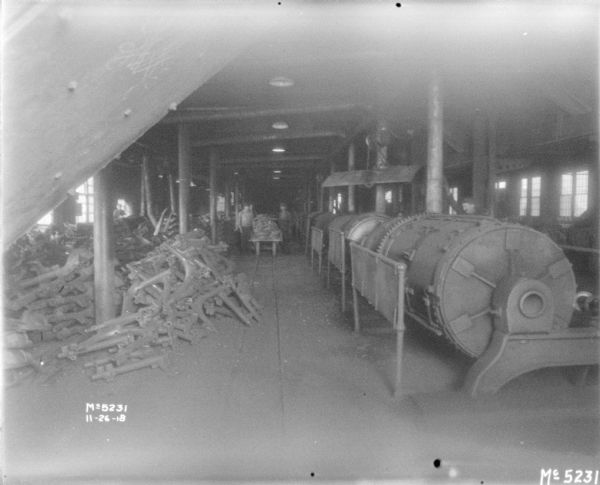 View of manufacturing area. There are parts stacked on the floor on the left, and machinery is on the right. Men are standing in the background near a cart stacked with parts.