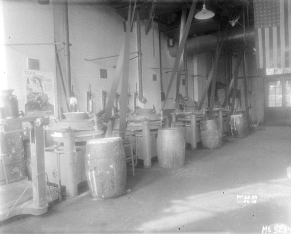 Four barrels are in front of four machines sitting indoors on a factory floor. The machinery is belt driven, and the belts are suspended from the ceiling. A large U.S. flag is hanging over a doorway in the background on the right. A poster on the wall on the left reads: "Team Work Wins!"