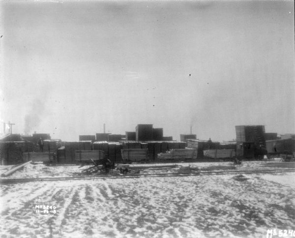 View across snowy yard towards stacks of lumber. Men are standing on carts loaded with lumber.