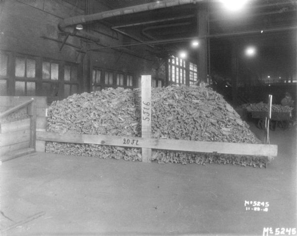 A pile of links are in a pile on a floor behind a wood fence, and a sign on the fence reads: "20 Ft." Another sign reads: "5 Ft 6." A man is standing near a cart in the background on the right.