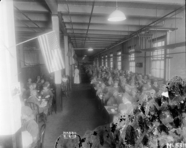 Slightly elevated view of women eating in the dining room. They are posing for the camera. Flags are hanging from the columns in the room.