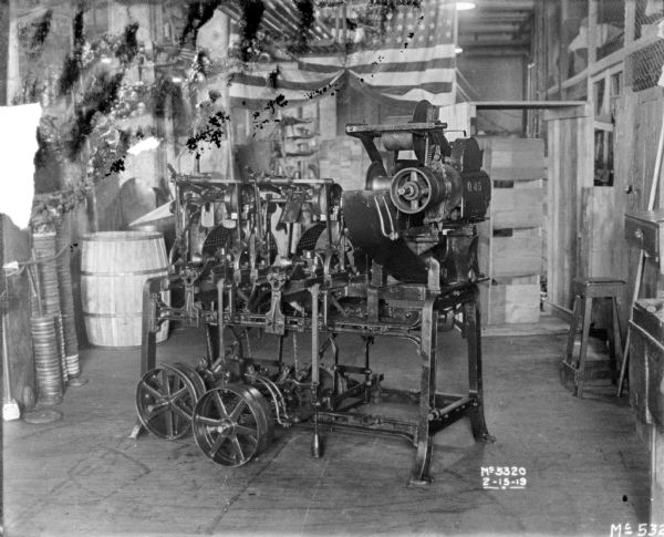 Machine on factory floor. There are parts and a barrel, and a U.S. flag in the background.