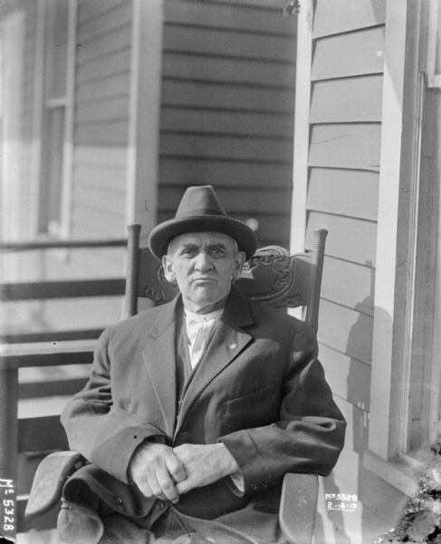 Elderly man, in dress clothes and hat, sitting in a rocking chair outdoors in front of a building.