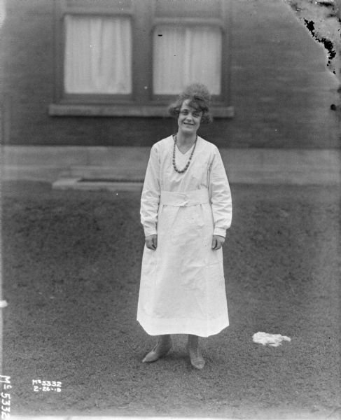 Full-length portrait of a young woman in a white dress standing outdoors. There is a building in the background.