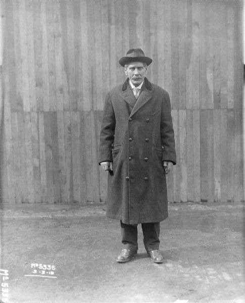 Full-length portrait of a man standing outdoors. A fence or building is in the background.