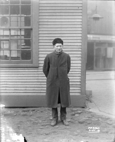 Full-length portrait of a man standing outdoors in front of a building. He is wearing a long coat and a hat.