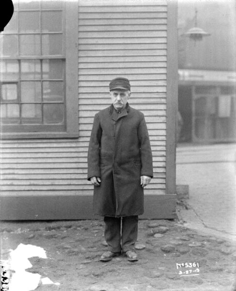 Full-length portrait of a man standing outdoors in front of a building. He is wearing a long coat and a hat.