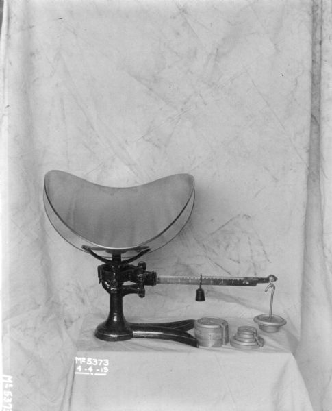 An 8 ounce scale is set up on a cloth covered stand, with a cloth backdrop behind. Additional weights are next to the base.