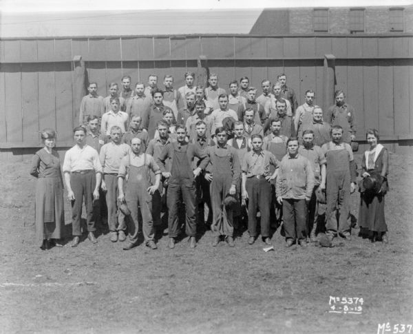 A large group of men, and two women, are standing outdoors in front of a fence. The women are wearing dresses, and the men are wearing work clothes.