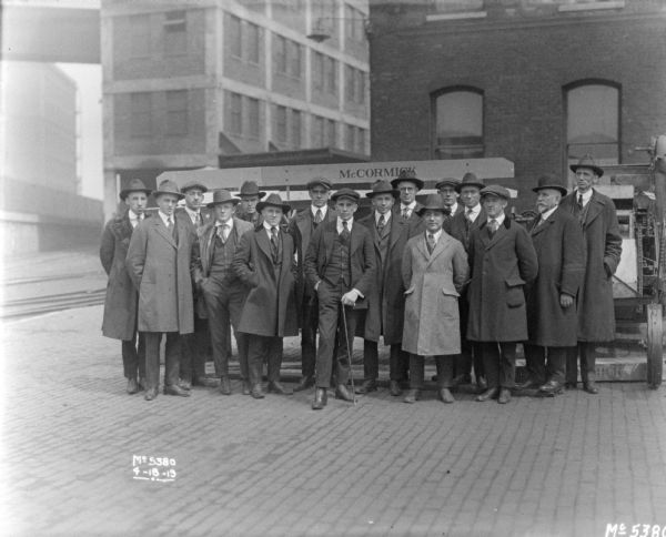 Group portrait of men wearing suits, hats and coats posing in front of a Harvester Thresher in factory yard. In the background are factory buildings.