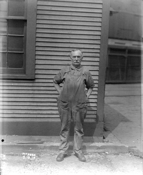 Full-length portrait of a man standing outdoors in front of a building. He is wearing work overalls.