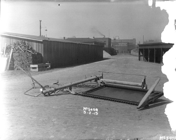 Binder part outdoors in factory yard. There is a long, low building in the background on the left, and an elevated railroad platform on the right. A sign on a building in the far background reads: "McCormick Station."