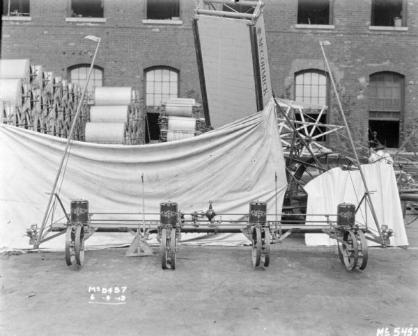 Planter outdoors in factory yard with a cloth backdrop behind it. The backdrop is attached to another agricultural machine, which has a section raised up in the air to hold the backdrop. In the background are large stacks of what may be parts, and a brick building. A man is working in the yard on the right.
