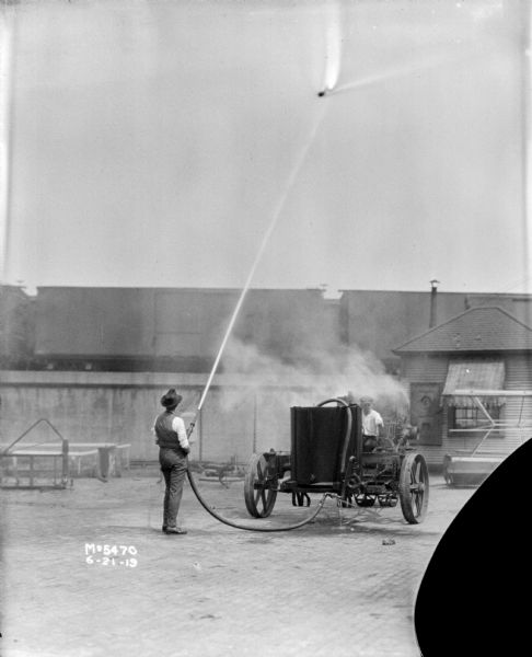 A man is standing in the factory yard using a sprayer, while another man is sitting on the machine with the tank. In the background, railroad cars are on an elevated railroad platform.