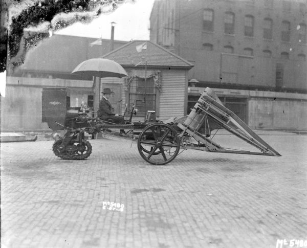 A man is sitting under an umbrella on a tractor-pushed Corn Picker in factory yard. Perhaps for research and development. There is a small building in the background near railroad cars on an elevated railroad platform.