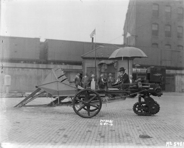 A man is sitting under an umbrella on a tractor-pushed Corn Picker in factory yard. Perhaps for research and development. A group of men are watching and standing in front of a small building in the background. Behind the building are railroad cars on an elevated railroad platform.