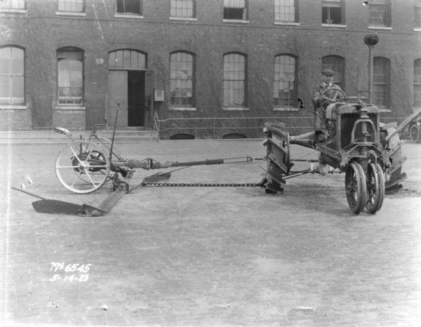 A man is sitting on a tractor pulling a mower in factory yard. A brick building is in the background.