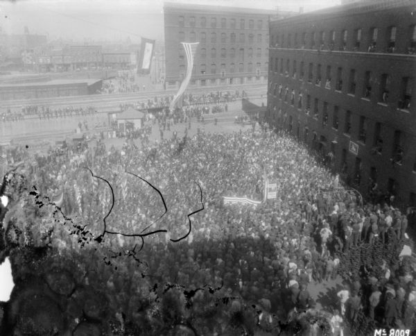 Elevated view of a rally of employees for liberty loan campaign. The factory yard is crowded with people, and other people are sitting on the elevated railroad platform. People are looking out of the windows of the brick factory building on the right. Banner and flags are handing from cables strung from the buildings.