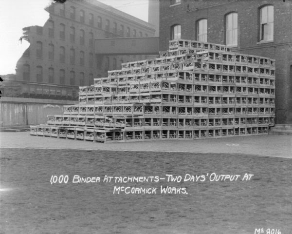 View of stacked crates holding Binders in factory yard. In the background are two brick buildings connected with a skybridge, and below are railroad cars on an elevated railroad platform. Text at bottom reads:  "1,000 Binder Attachments — Two days' output at McCormick Works."