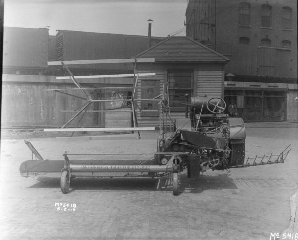 A tractor-drawn Binder in factory yard. A man is standing near a small building in the background, and above are railroad cars on an elevated railroad platform.