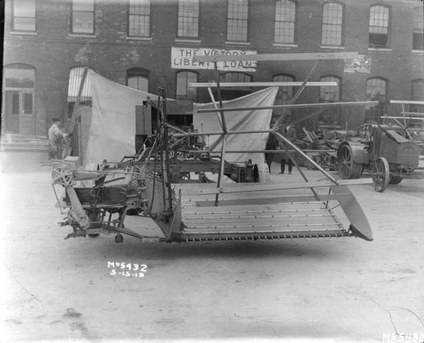 A tractor-drawn Binder in factory yard. A man is holding a piece of wood that is attached to the end of cloth backdrop that is set up behind the Binder. Other men are standing on the right near a brick building. A banner on the factory building reads: "The Victory Liberty Loan."