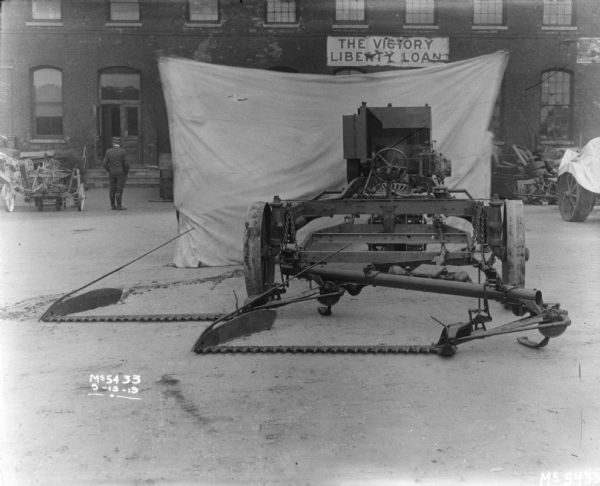 A tractor-drawn Binder in factory yard with a backdrop behind it. A man is standing in the background near a brick building. A banner on the factory building reads: "The Victory Liberty Loan."