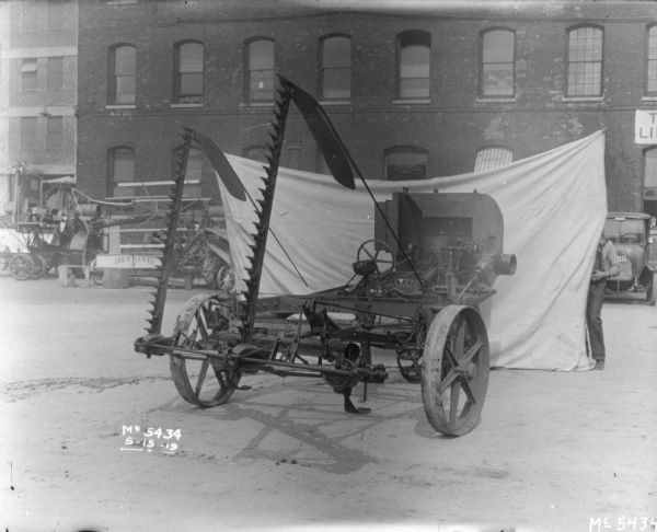 A tractor-drawn Binder in factory yard. Men are holding cloth backdrop that is set up behind the Binder. There is a banner on the factory building, which probably reads: "The Victory Liberty Loan."