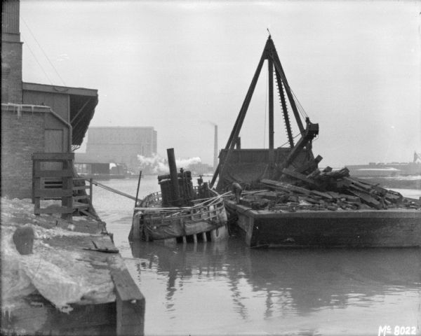 View from shoreline towards a barge alongside a boat. A man is standing on the barge. There are buildings in the background.