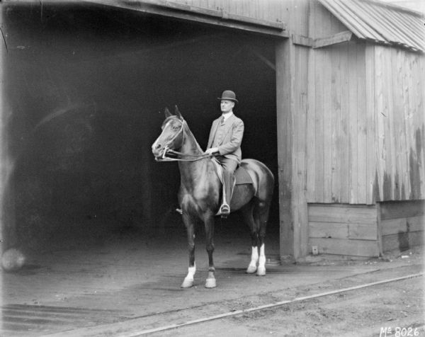 Man on horseback near a barn. He is wearing a suit and hat.