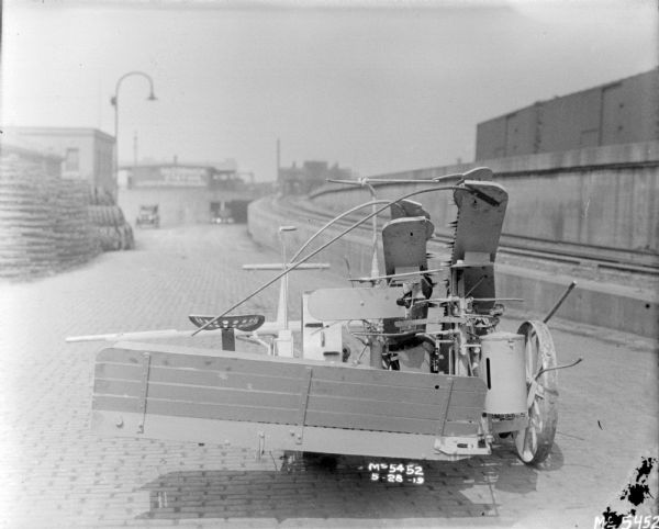 Rear view of a Corn Binder in factory yard. Agricultural machinery and parts are in the yard in the background. A sign on a building in the distance reads: "McCormick Station." On the right is an elevated railroad station.