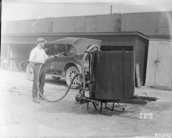 Man filling gas tank of car from gas tank mounted on sled. In the background is an open-sided shed and railroad cars on an elevated railroad platform. 