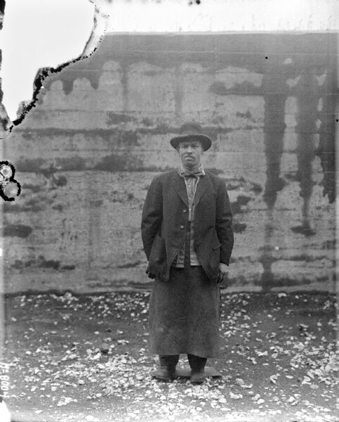 Full-length portrait of a man standing outdoors in front of a wall. He is wearing a hat, jacket and long apron.