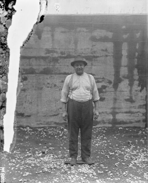 Full-length portrait of a man standing outdoors in front of a wall. He is wearing a hat, shirt and pants.